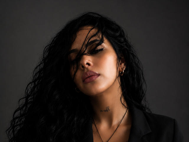 NattParis has dark wavy hair covering one eye, brown skin, gold earrings, and a small tattoo below her chin. She's wearing a black jacket with a subtle, sexy look.