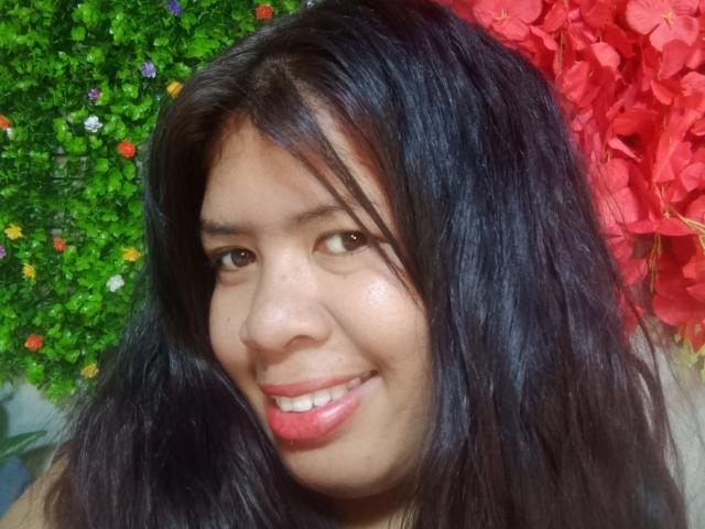 MariahSquirting69, con la pelle marrone chiaro, i capelli neri ondulati, gli occhi marroni e un leggero sorriso che mostra il rossetto rosa, sullo sfondo di foglie verdi e rosse..