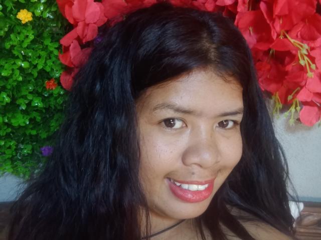 MariahSquirting69 has medium brown skin, black wavy hair, red lipstick, and a slight smile. Red and green floral background. Small yellow, purple, and red flowers on left.