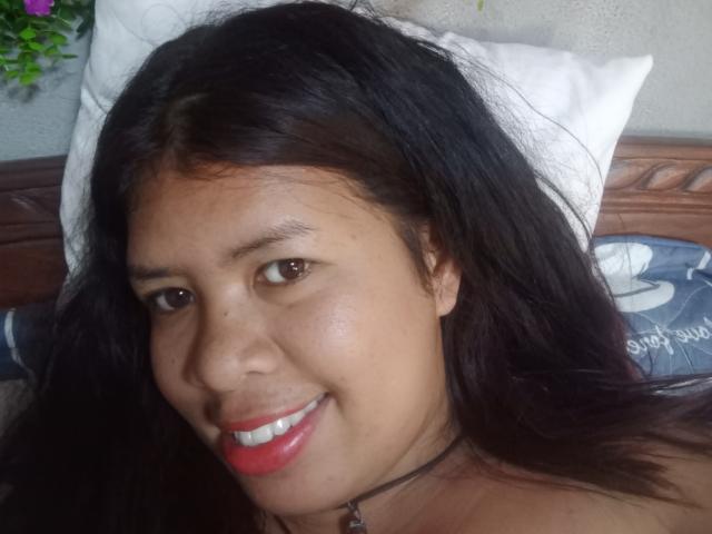 MariahSquirting69 with long black hair, brown eyes, and red lipstick, smiling at the camera. Her small tits and dark brown skin are visible. She's wearing a black choker.