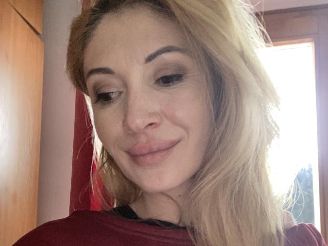 RebelLuna has blonde hair, slightly wavy, with dark eyebrows and a subtle smile. She's wearing a red shirt, looking down with a cute, confident expression.