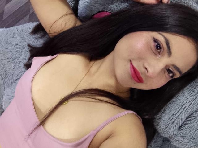 AngelAlba with long black hair, pink tank top, and red lipstick, lying on a grey blanket, showing her tits and smiling at the camera.
