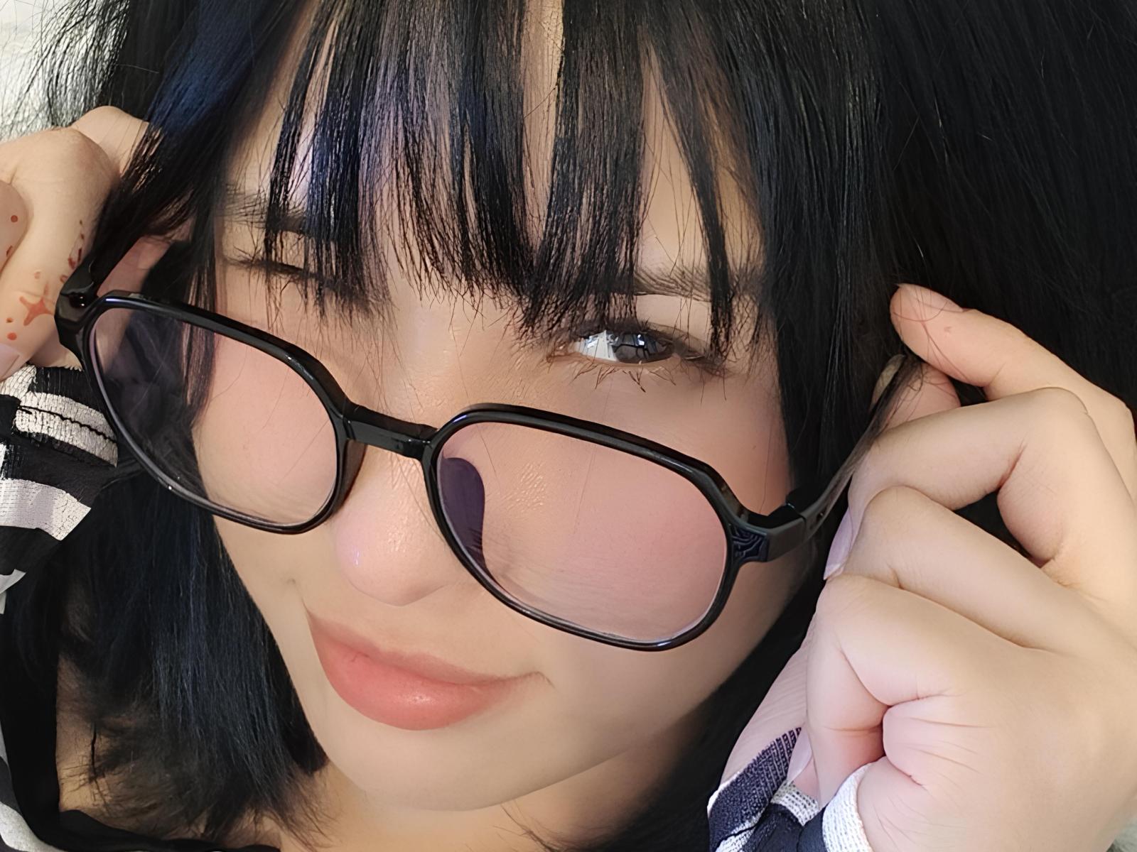 Chantell Houlihan, con gafas negras, sosteniéndolas con ambas manos, tiene el cabello negro y liso con flequillo, piel clara y lápiz labial rosa. Su pequeña y linda nariz está ligeramente levantada..