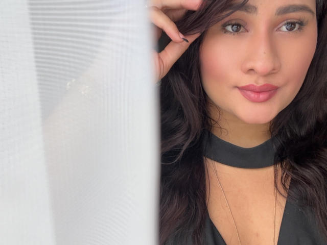 ElyRoss has brown wavy hair, a black choker, and pink lipstick. She's looking at the camera with a slight smile, her hand in her hair. Her tits are not visible.