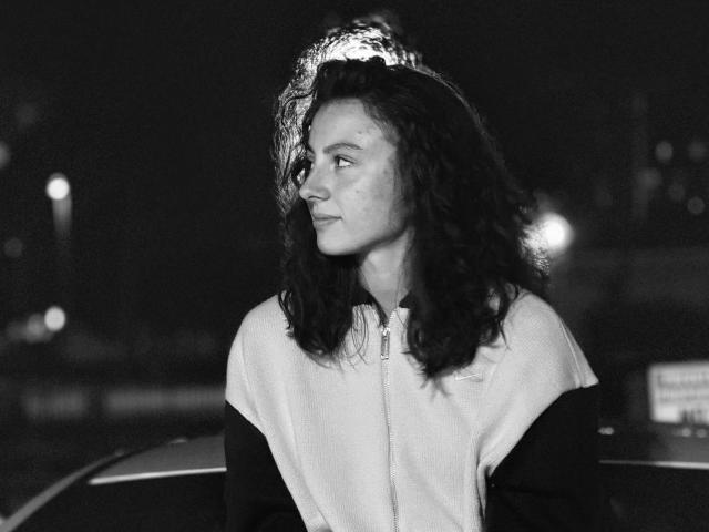 Black and white photo of WillaBelz with curly dark hair, light skin, and a serious expression, wearing a zip-up jacket, standing next to a car at night.