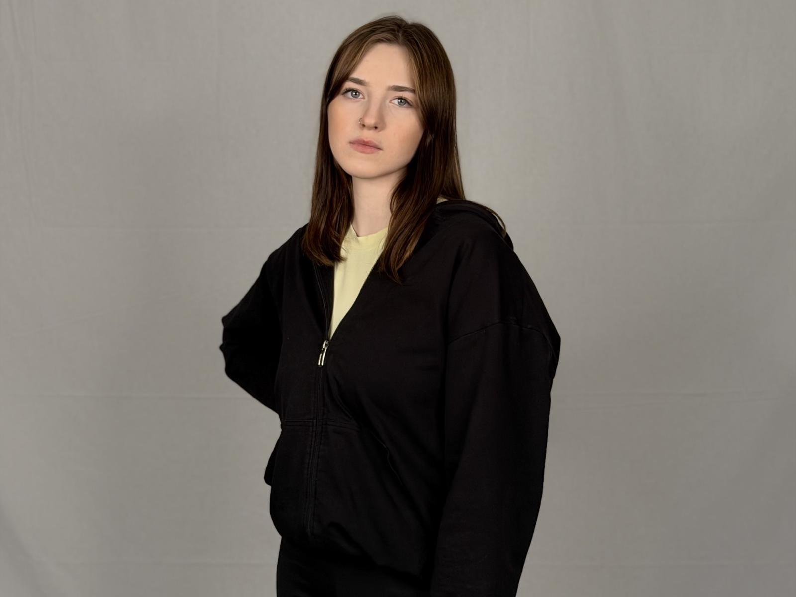 TrinhGanin has straight brown hair, pale skin, and blue eyes. She wears a black zip-up jacket over a yellow shirt, standing with a neutral, slightly serious expression.