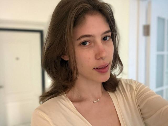 Terrilyn Lessey mit hellbraunen Haaren, Nasenring und einer "Liebe"-Kette, trägt ein beiges Oberteil und blickt mit einem leichten Lächeln in die Kamera, ihr Gesicht ist weich beleuchtet..