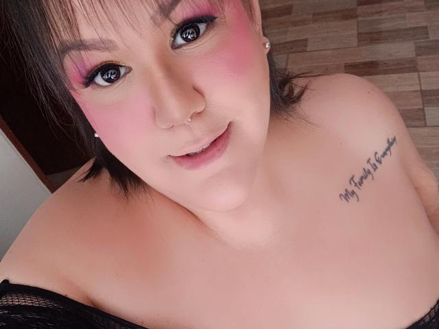 GiiannaMorrys has pink eyeshadow, a nose ring, and a tattoo on her right shoulder. She's smiling with bare shoulders, wearing black lace. Her tits are partially visible.