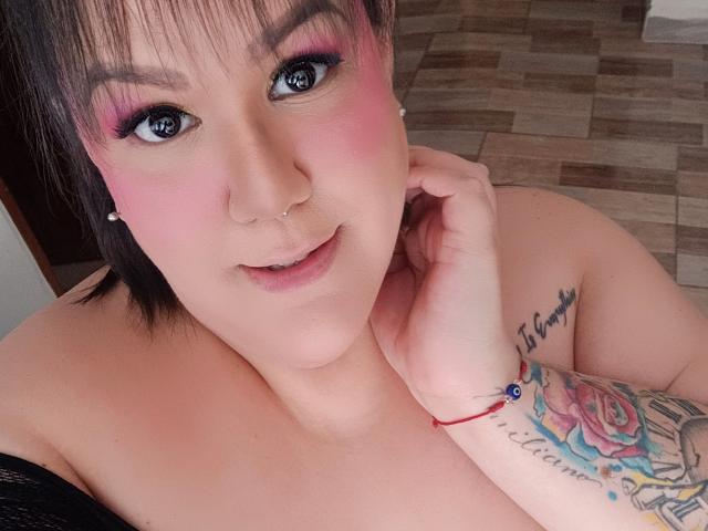 GiiannaMorrys with pink eyeshadow, blue eyes, short black hair, fair skin, small nose ring, smiling, hand on neck, tattooed arm, red bracelet, and visible bare breasts.
