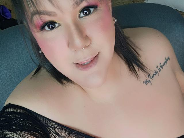 GiiannaMorrys has pink eyeshadow, a nose piercing, and black eyeliner. She's smiling with exposed shoulders and a tattoo on her right chest. She's wearing a black lace top.