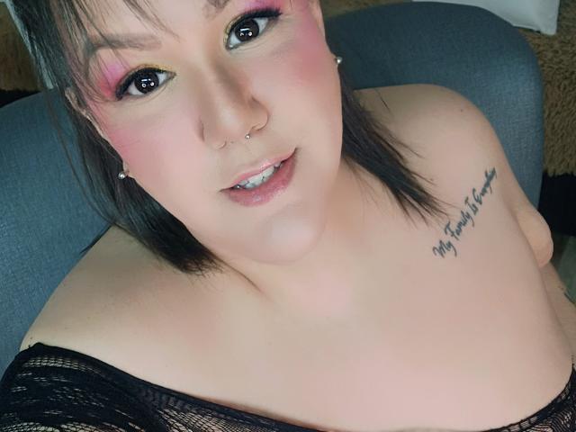 GiiannaMorrys with pink eyeshadow, black nose ring, smiling, showing bare shoulders, black lace top, tattoo on right shoulder, short black hair, looking up. Boobs visible.