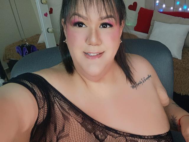 GiiannaMorrys with pink eyeshadow, black mesh top, showing tits, small nose ring, tattoo on right shoulder, smiling, dark hair, fair skin, seated in chair, arm raised.