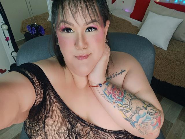 GiiannaMorrys with pink eye makeup, black lace top, showing her tits, tattooed arm, nose ring, and pink lips, holding her cheek, sitting in a chair, smiling.