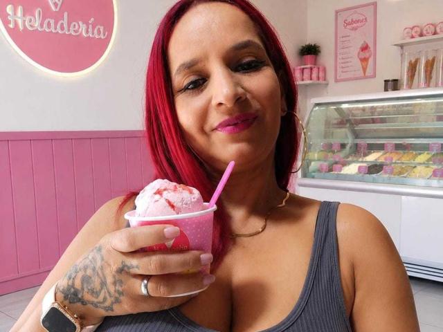 KateMonroe with red hair, pink lipstick, and tattoos, holding a pink ice cream with a pink straw, smiling at the camera, wearing a gray tank top, in a pink-themed ice cream shop.