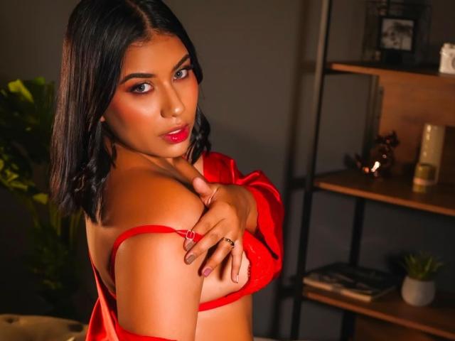 Mia Cameron, com cabelo escuro, batom vermelho e sutiã vermelho, cobrindo seus pequenos seios com uma mão, olhando por cima do ombro, em um quarto com pouca luz..