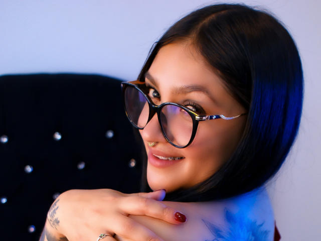 MiaaVixen with black glasses, nose ring, and red nails, smiling at the camera, holding her shoulder with tattooed hand, blue light on right side of her face.
