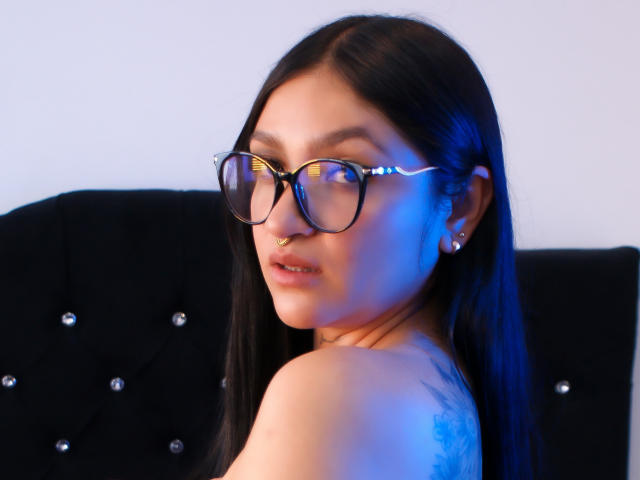 MiaaVixen with long black hair, glasses, and a nose ring, looking over her shoulder, showing a tattoo on her upper arm, in soft blue and white lighting.