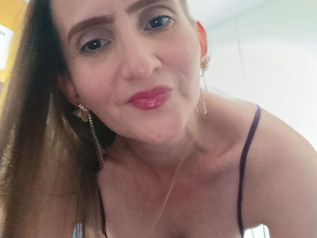 Rubi Anderson, con rossetto rosa, occhi azzurri e capelli castani, indossa una canottiera viola, orecchini e una collana, e sorride leggermente con le labbra leggermente arricciate..