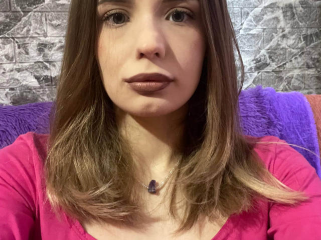 JoyeWakefield, com cabelo castanho, camisa rosa, colar roxo e batom roxo, olhando diretamente para a câmera. Seus seios são pequenos e ela tem um rosto bonito..
