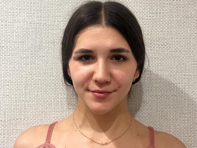 MiguelinaValenstein has dark brown hair, fair skin, and is wearing pink lace straps with a thin gold necklace, smiling gently at the camera with small, subtle pink lips.