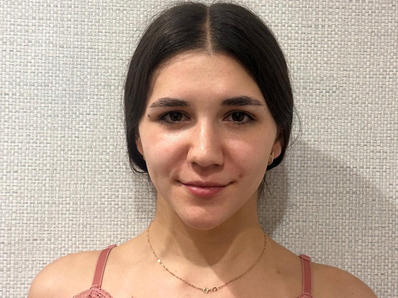 Miguelina Valenstein tiene el cabello castaño oscuro, piel clara y lleva tirantes de encaje rosa con un delgado collar dorado, sonriendo suavemente a la cámara con labios pequeños y rosados..