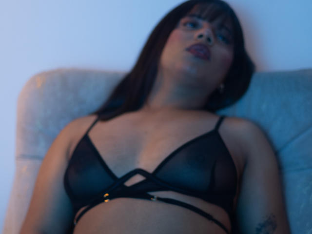 ZaraBlaze is lying back, wearing a sheer black bra with gold rings, showing her tits, with dark brown hair and red lipstick, in a slightly blurry, blue-lit photo.