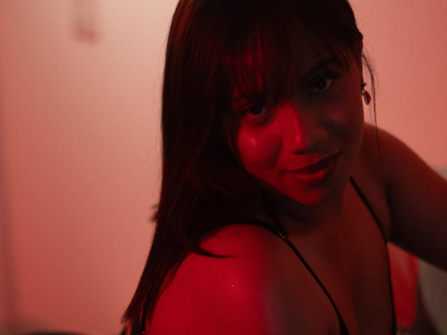 ZaraBlaze has red-lit skin, brown hair, and a slight smile. She's wearing a black spaghetti strap top, showing her small tits and a glistening shoulder.