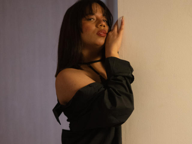 ZaraBlaze with dark straight hair, black off-shoulder top, and red lipstick, kissing her hand, showing bare shoulder and collarbone, standing against a white wall.