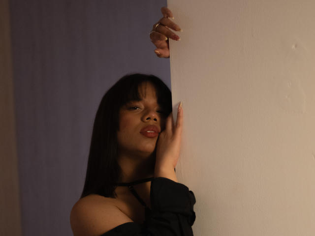 ZaraBlaze with dark straight hair, pouty lips, and black off-shoulder top, hands on white wall, right hand gripping edge, left hand touching wall, subtle sexy look.