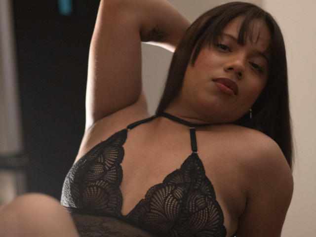 ZaraBlaze with medium brown skin, straight brown hair, and red lipstick, wearing a black lace bra, arm raised, showing small tattoo on her bicep, and slightly parted red lips.
