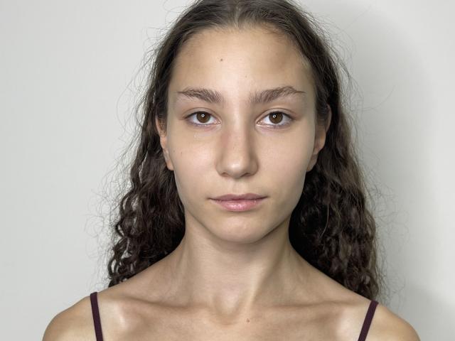 ShantaeTsuha has brown eyes, wavy dark brown hair, and a neutral expression with slightly parted pink lips. She has a slender neck and wears a dark purple spaghetti strap.