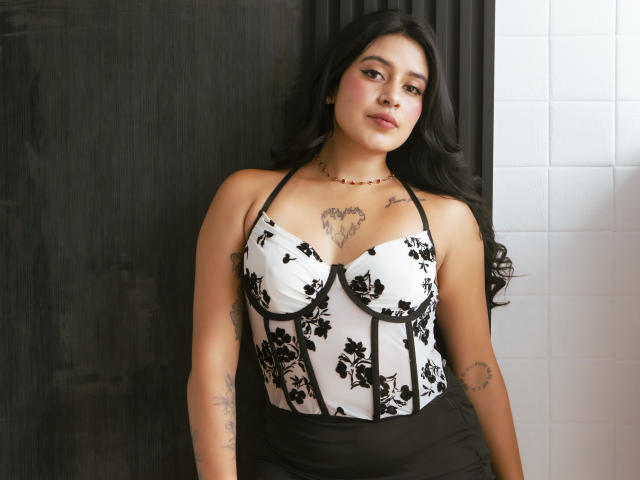 MillaDurand with medium tits in a black and white floral halter top, black skirt, tattoos on chest and arms, long black hair, subtle makeup, standing against dark and white.