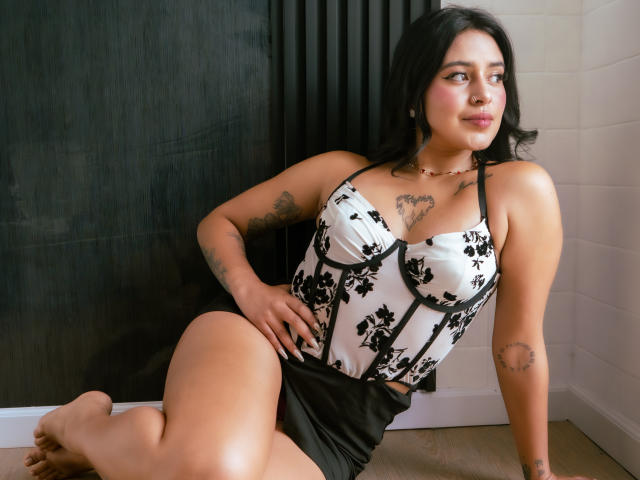 MillaDurand with black hair, wearing a white and black floral corset, sitting with one leg up, showing tattoos, and looking to the side with a slight smile.