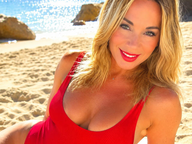 EllisaWoman with blonde hair, red lipstick, and red tank top showing big tits, sitting on sunny beach, smiling at camera, blue ocean and rocks in background.