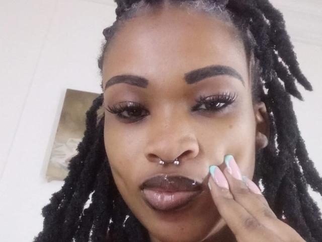JuiceLipsx with dark brown skin, black dreadlocks, nose piercings, and glossy lips, tongue slightly out, holding her chin with pastel-colored nails, wearing thick eyelashes.