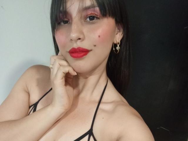 MadissonFontaine with dark hair, red lipstick, and pink eyeshadow, wearing a black strappy top, hand on chin, small moles on her cheek, gold heart earrings, and a ring.