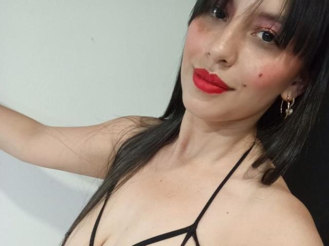 MadissonFontaine, avec des lèvres rouges, un haut noir à bretelles, une peau pâle, de petites taches de rousseur sur le visage, des cheveux noirs, des boucles d'oreilles, un léger sourire, une décolleté visible, un éclairage doux, la tête légèrement inclinée..