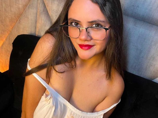 Emily Colsson, met een bril, rode lippenstift en een witte jurk die haar grote borsten laat zien, zittend op een zwarte bank, lachend naar de camera, bruin haar, lichte huid..
