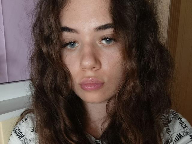 Hannelore Bucknell heeft golvend bruin haar, blauwe ogen en glanzende roze lippen, met een lichtjes fronsende uitdrukking, en draagt een witte blouse met zwarte tekst..