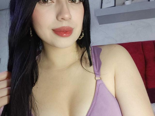 Isabella Rossi, met lang zwart haar, lichte huid, roze lippenstift, draagt een paars tanktop, waarbij haar kleine borsten zichtbaar zijn, gouden oorbellen en ze heeft een lichte glimlach op een selfie..