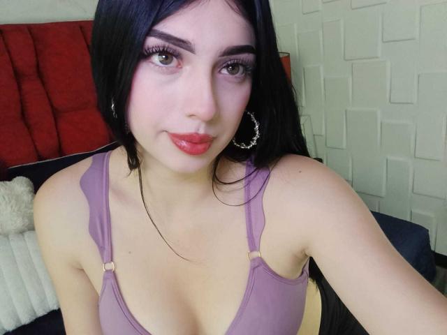 Isabella Rossi, met lang zwart haar, een paarse bh en roze lippenstift, die haar borsten laat zien, met oorringen en die de camera aankijkt met een lichte glimlach..