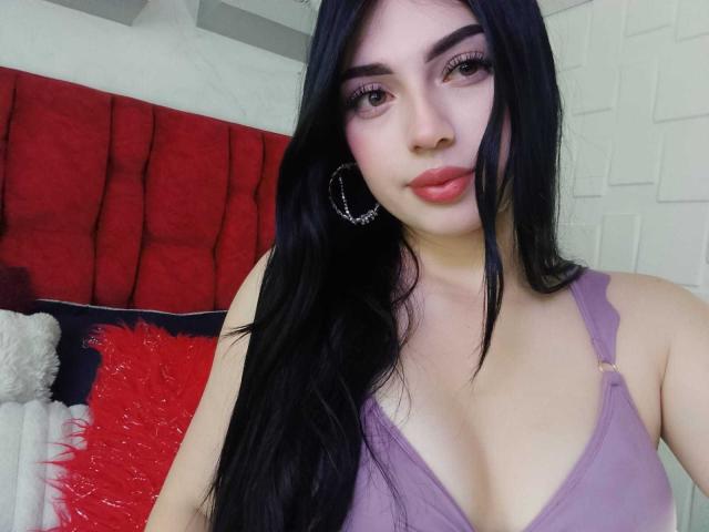 IsabellaaRossi with long black hair, purple bra, hoop earrings, and pink lipstick, showing her boobs, looking at the camera with a slight smile.