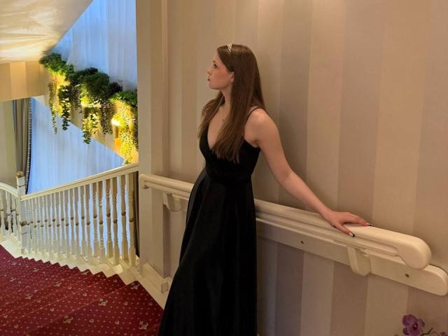 MelisaMagnie with long brown hair, wearing a black dress, standing against a white railing, looking to the left, in a carpeted hallway with green garland.