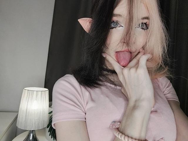 ChangCaddle with pink and black hair, pink cat ears, blue eyes, tongue out, hand near mouth, wearing pink top, white bracelet, lamp on left, dark background.