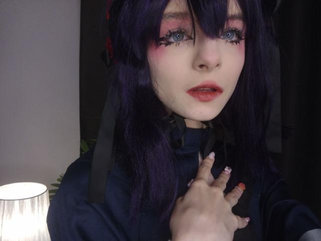 ChangCaddle with purple hair, blue eyes, pink eyeshadow, red lips, black ribbon, and dark shirt, touching her chest with painted nails, under a lit lamp.
