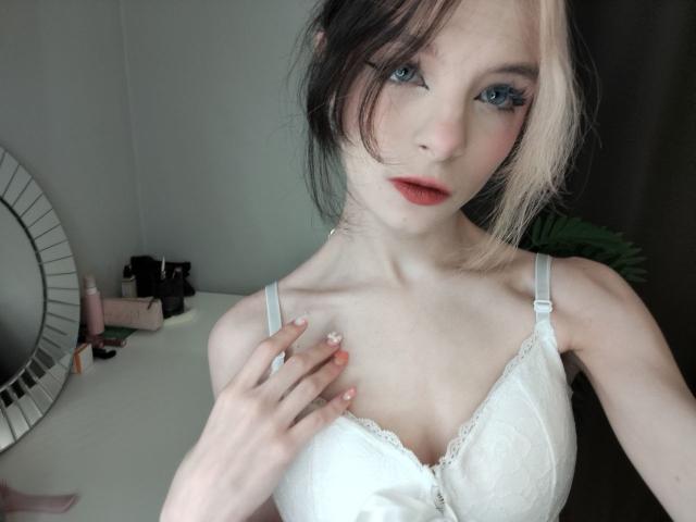 ChangCaddle with pale skin, blue eyes, and pink-black hair, wearing a white lace bra, touching her chest with painted nails, looking at the camera with red lips.
