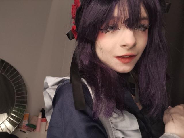 ChangCaddle with purple hair, red lipstick, and black eyeliner, wearing a dark blue and white outfit, showing a cute, slightly mischievous smile, with a red flower in hair.