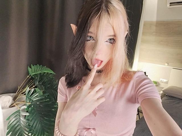ChangCaddle with split black and blonde hair, elf ears, pink shirt, finger in mouth, looking at camera, pale skin, small tits, cute, bedroom, potted plant, soft lighting.