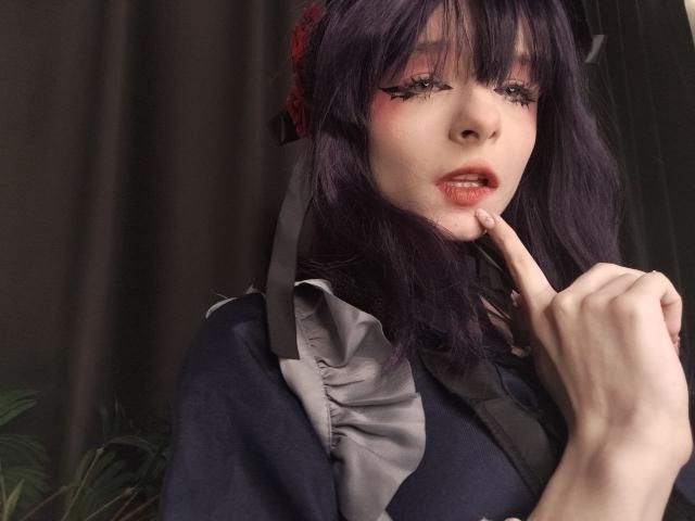 ChangCaddle has dark purple hair, red eyeliner, and pink lipstick, with a finger near her slightly open mouth. She's wearing a dark blue dress with a gray ruffle.
