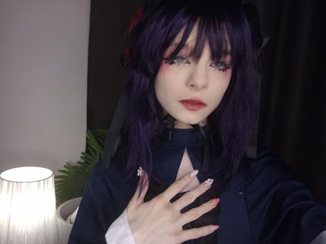 ChangCaddle has dark purple hair, pale skin, and red lipstick. She's wearing a black outfit with a ribbon, and her hand touches her chest, showing pink nails.